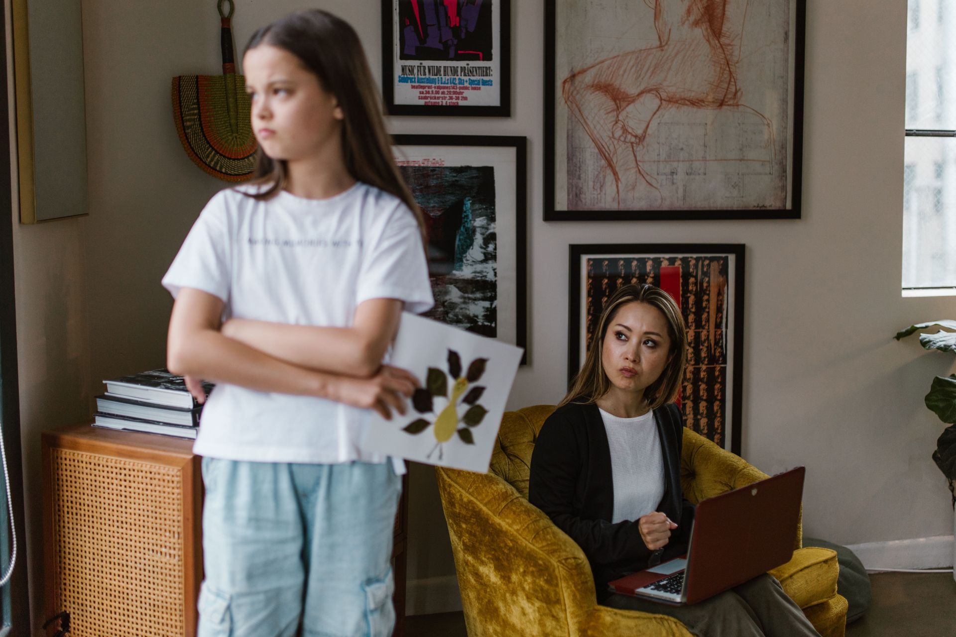 A mom and daughter in a bright living room, one standing angrily with an art project and one seated in a yellow chair using a laptop.