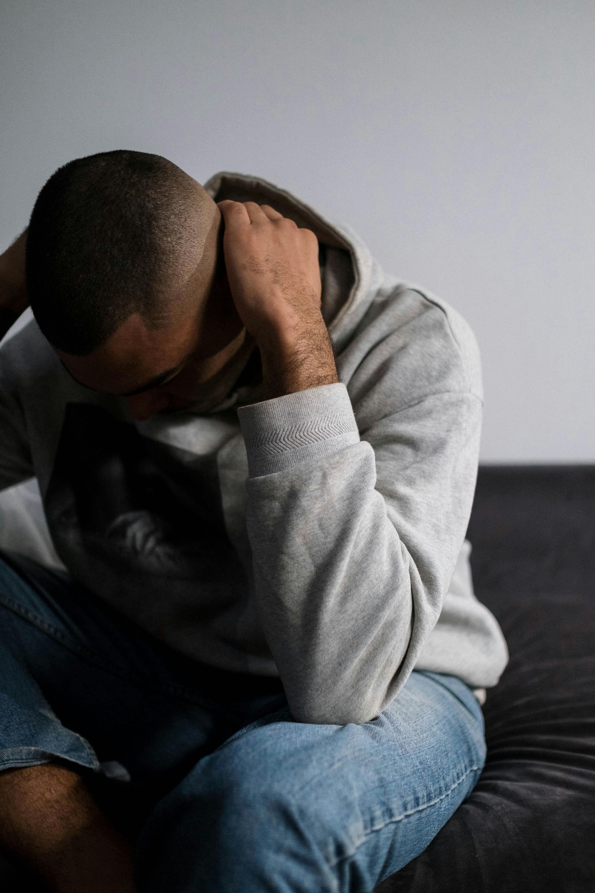 Person in a gray hoodie sits hunched on a bed, holding their head in distress.