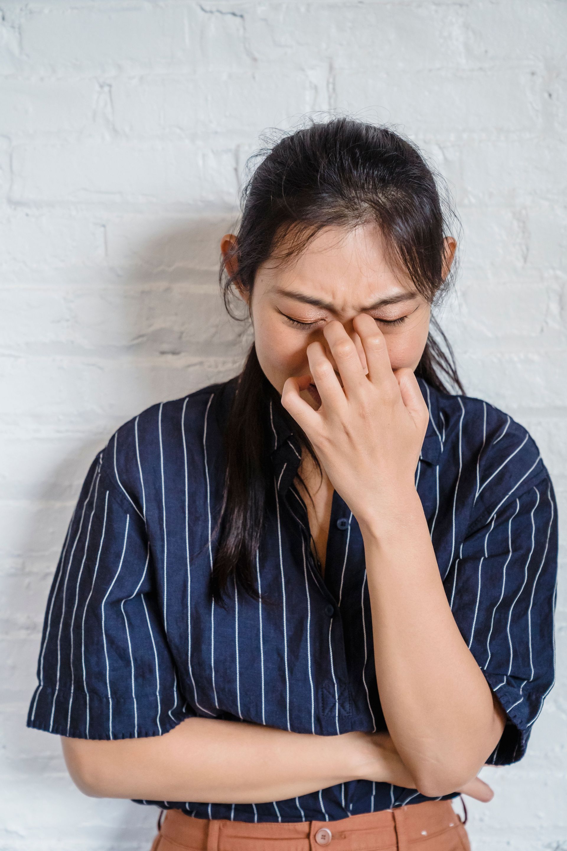 Person in a blue striped shirt covering their face with one hand against a white brick wall.
