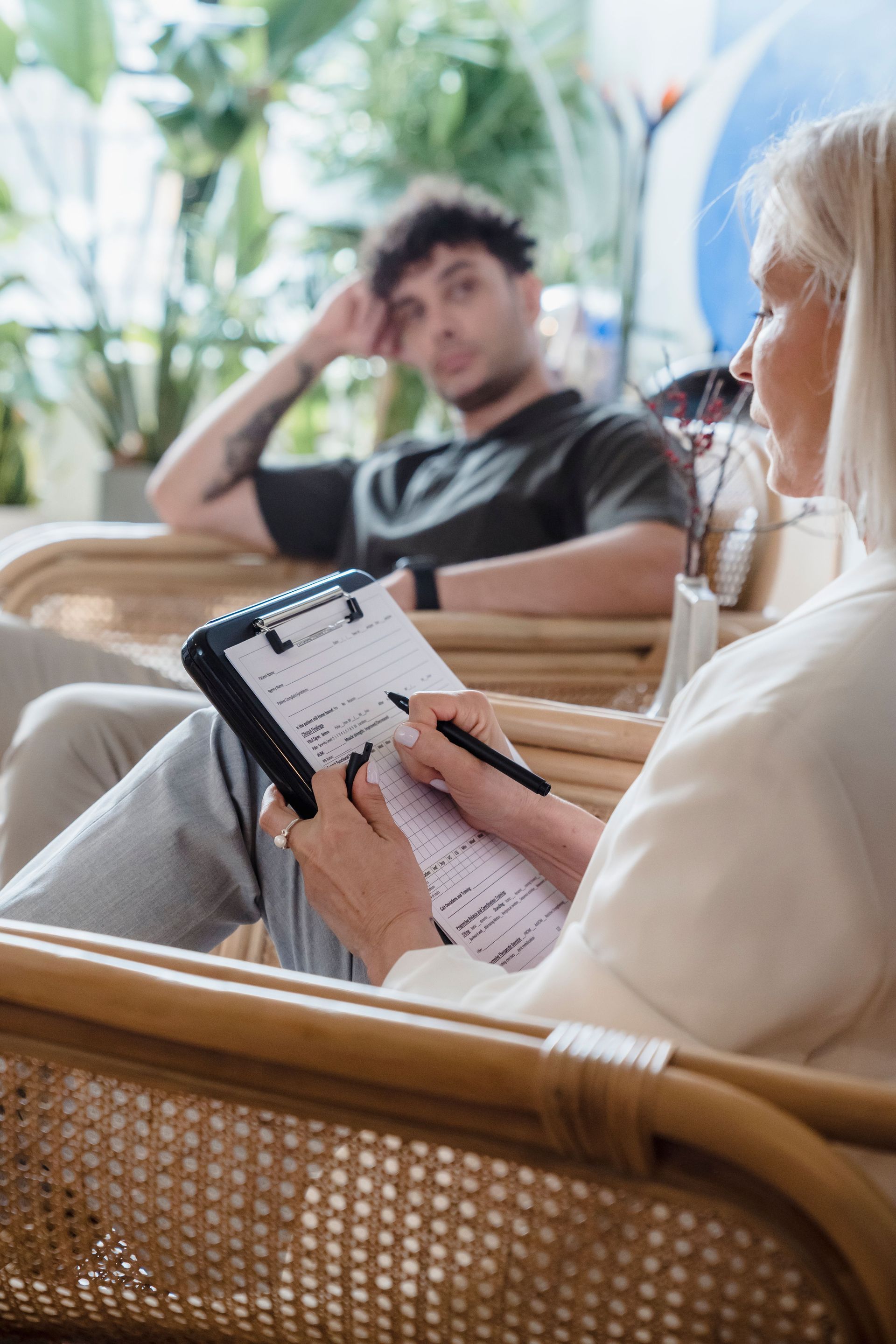 Two people sitting in wicker chairs, one writing in a notebook in a bright indoor lounge.
