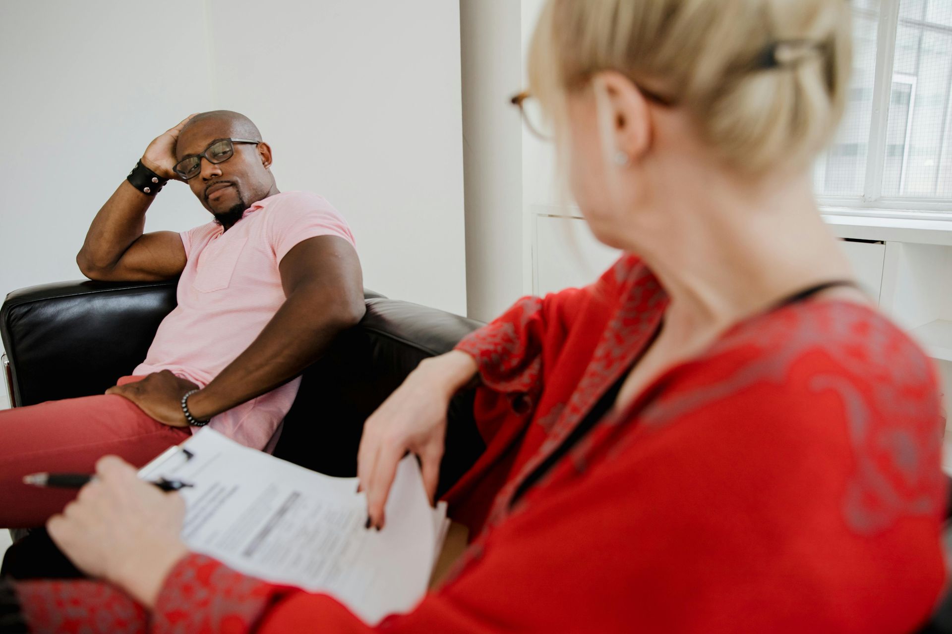 A Black man in a pink shirt sits on a couch and talks to a therapist in a red top holding a document.