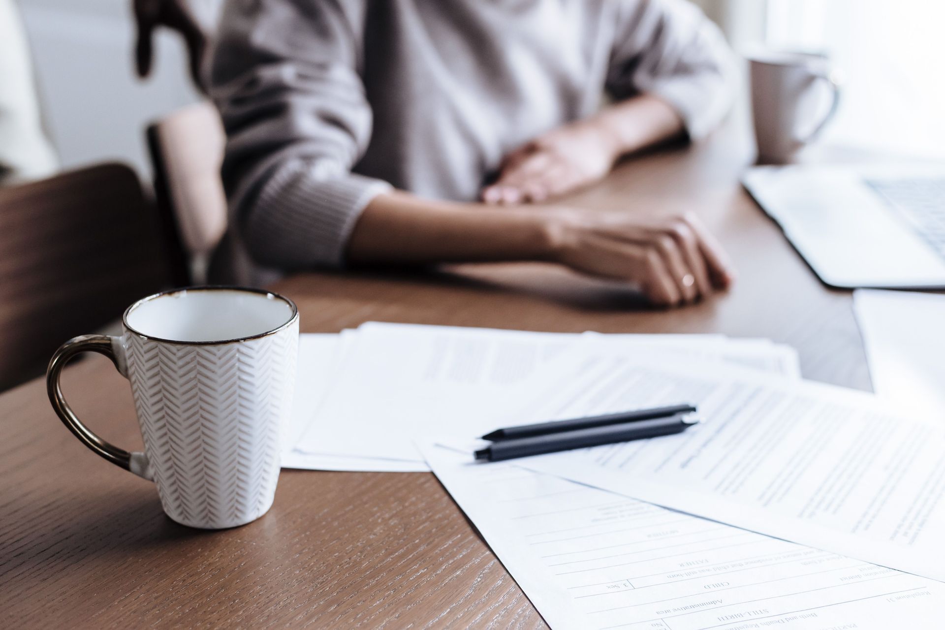 Coffee mug, pens, and paperwork on a table beside a seated person in a meeting room.