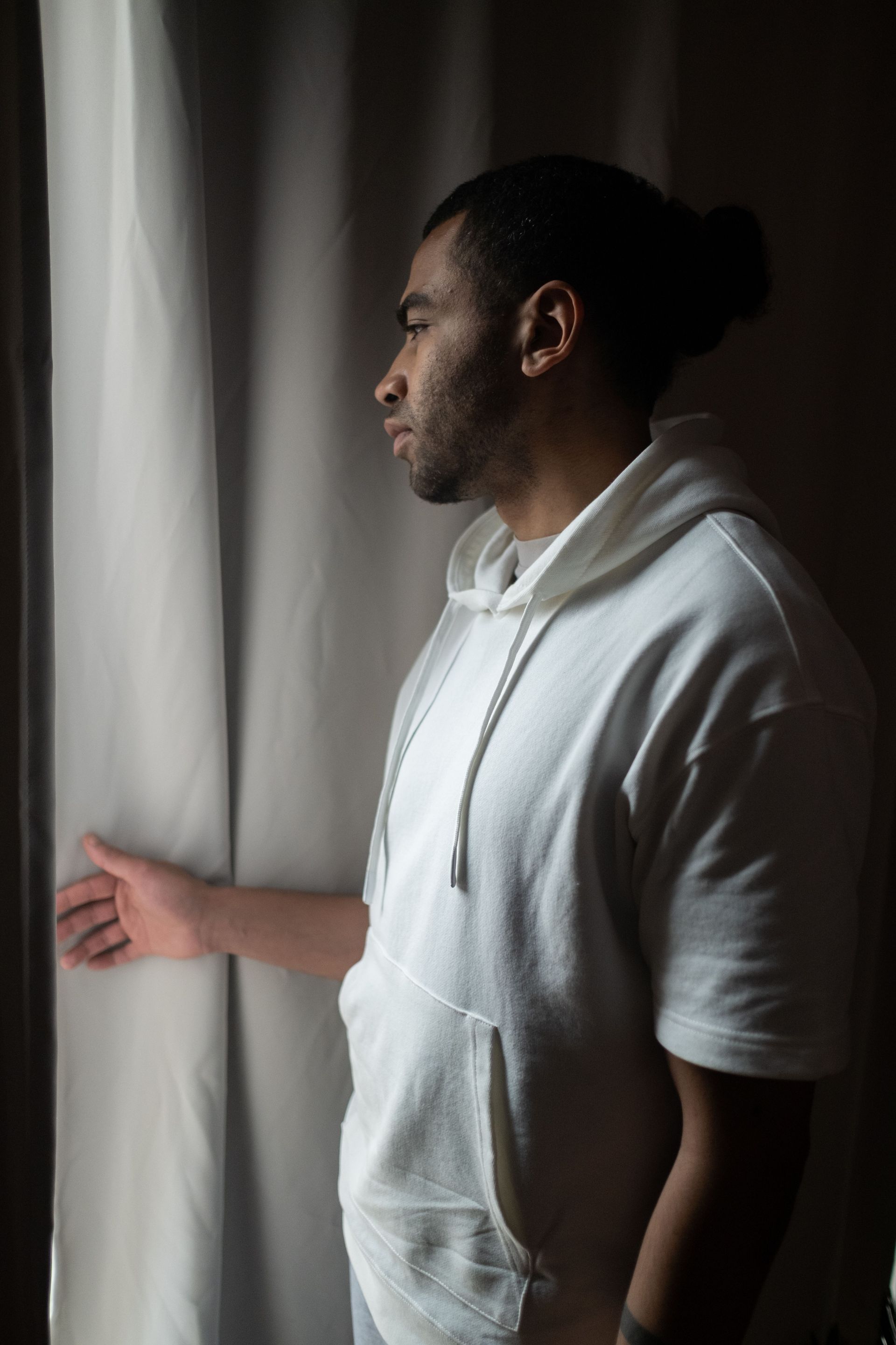 Person in a white hoodie standing by a window, looking outside in soft light.