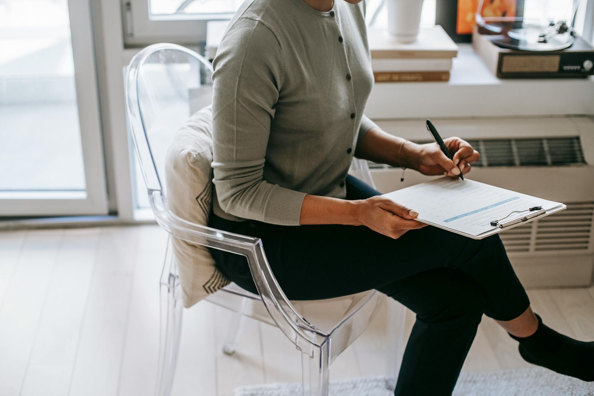Woman seated in a clear chair, writing on a clipboard in a bright modern room