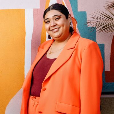 Woman in orange suit smiles, standing against colorful background.