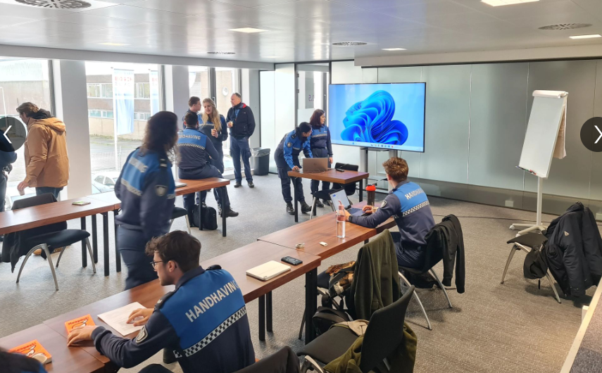 Mensen in blauwe uniformen in een vergaderruimte, sommigen zittend, sommigen staand, met een scherm waarop een afbeelding wordt getoond.