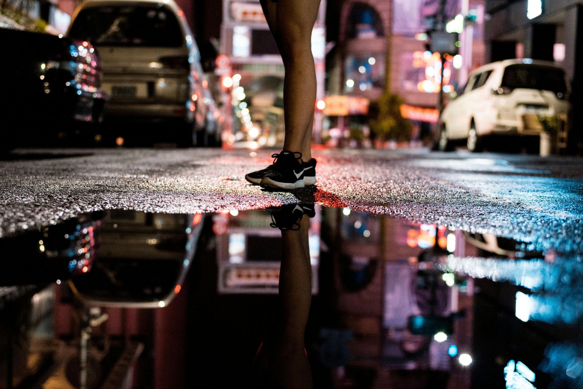 Een persoon staat 's nachts in een plas op een natte straat; de reflecties van auto's en stadslichten zijn zichtbaar.