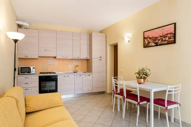 Piccola cucina e zona pranzo con mobili beige, tavolo e sedie bianchi e un divano giallo.