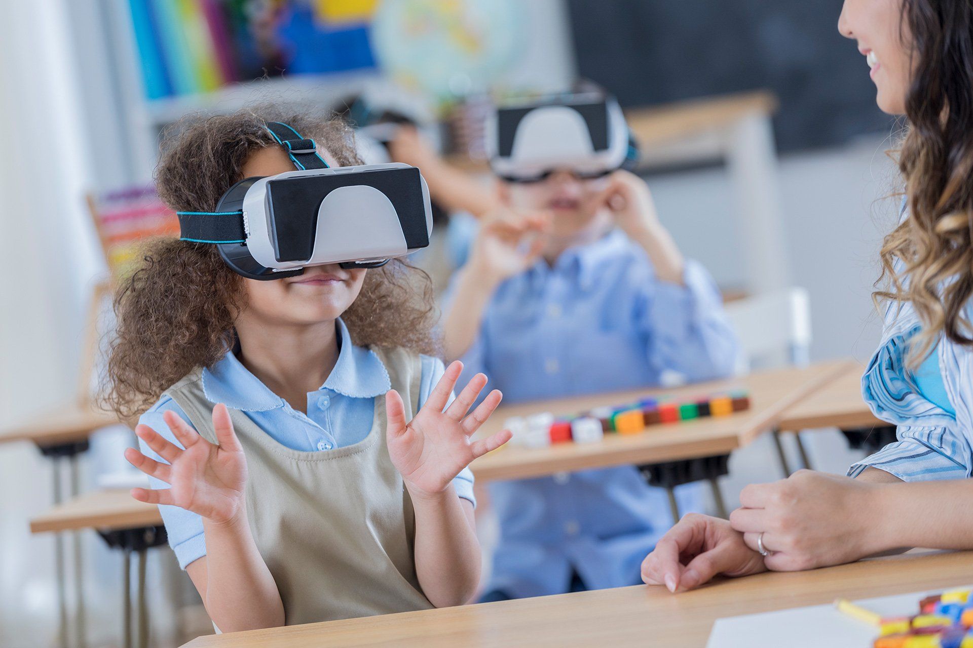 Girl wearing VR headset in classroom, gesturing with hands; teacher nearby, other student in background.