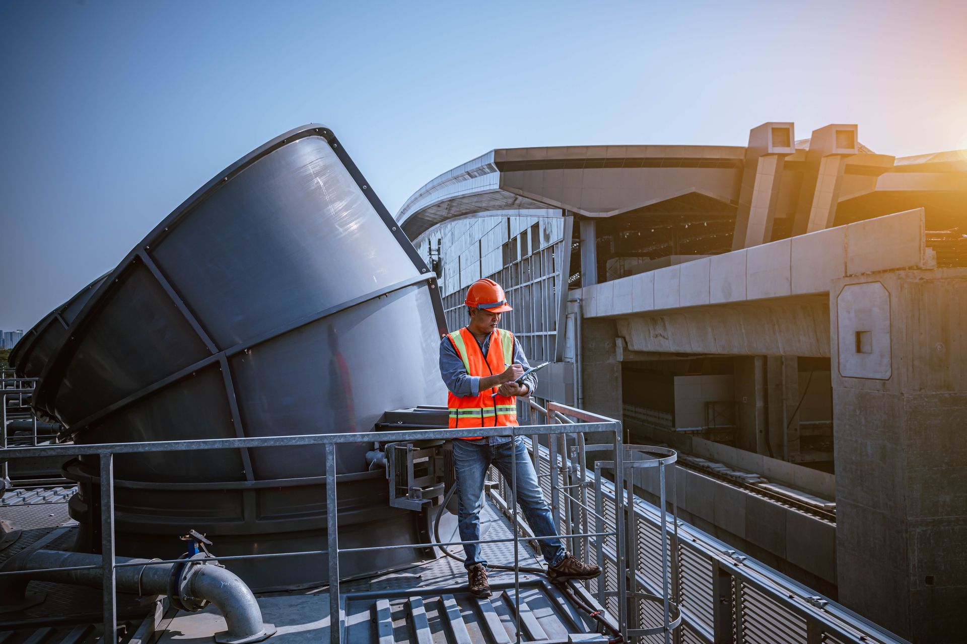 Worker in orange safety gear standing on a rooftop beside industrial machinery at sunset