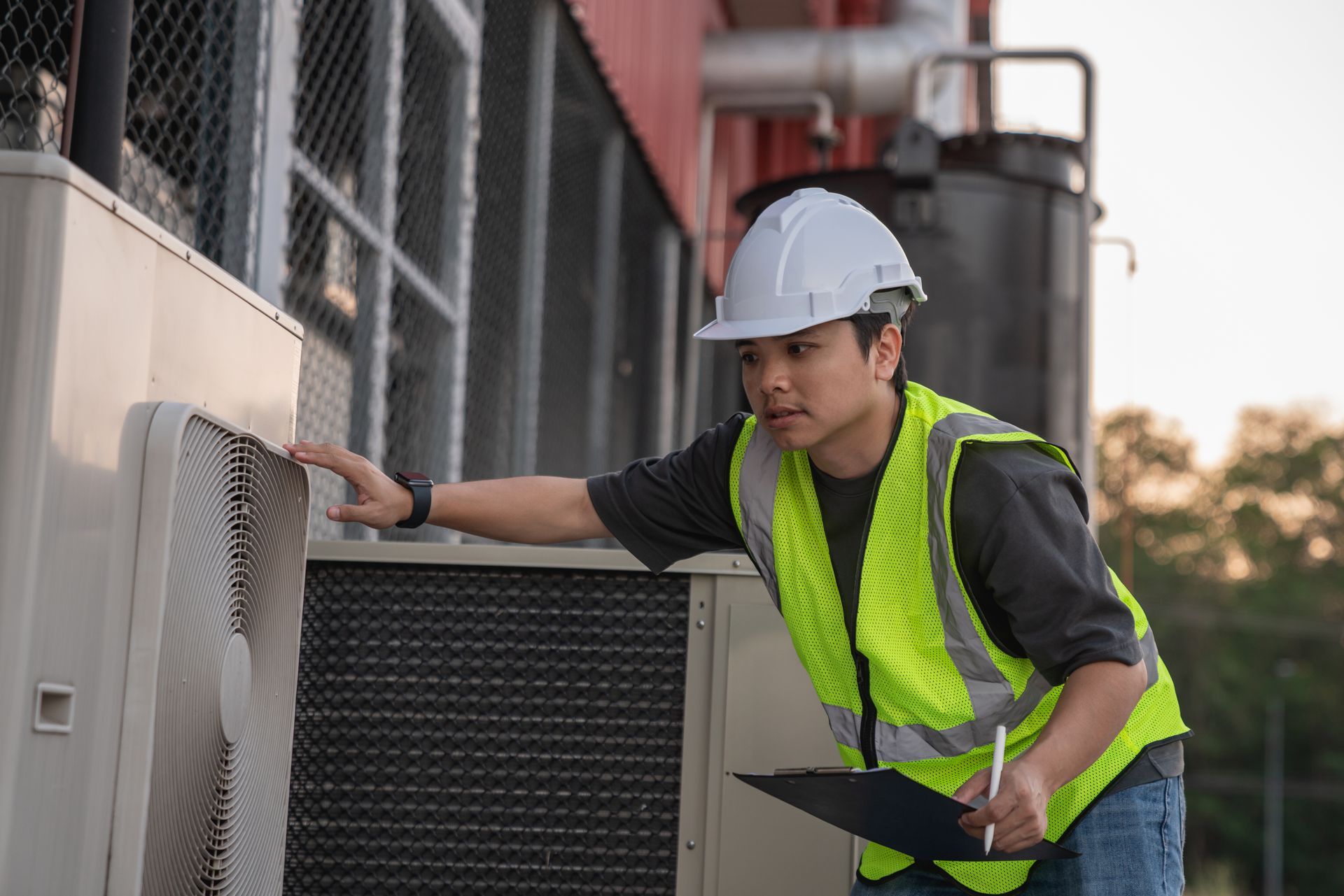 Worker in a hard hat and safety vest inspecting outdoor industrial equipment.