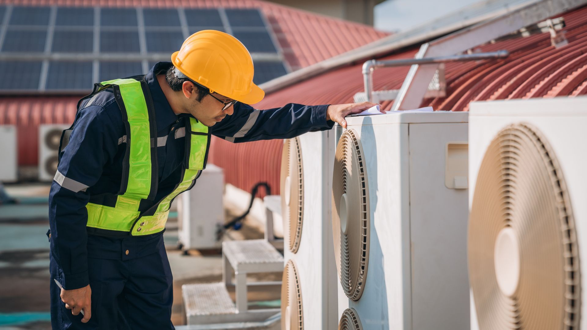 Worker in yellow hard hat inspects large rooftop HVAC equipment beside solar panels.