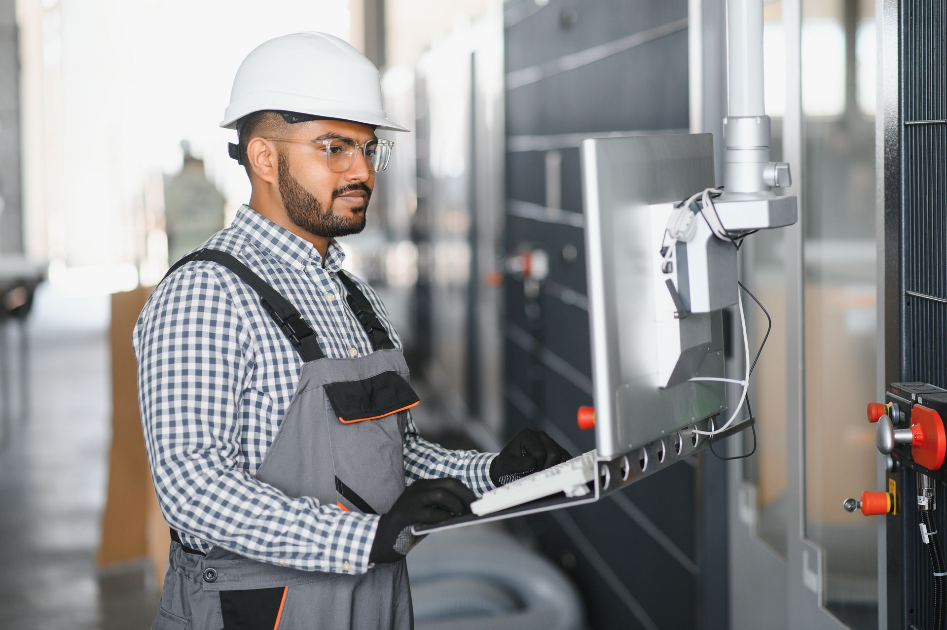 Technician in hard hat using a laptop and inspecting industrial equipment in a factory hallway
