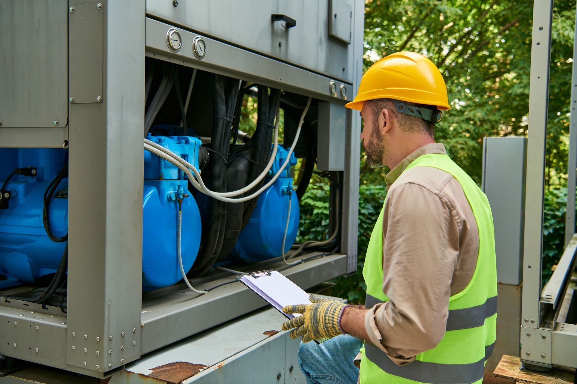 Technician in hard hat inspecting blue machinery outdoors with a clipboard