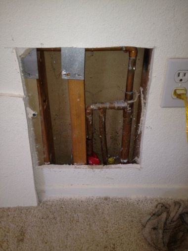 Wall cutout exposing studs and pipes beside an electrical outlet, with debris on the floor.
