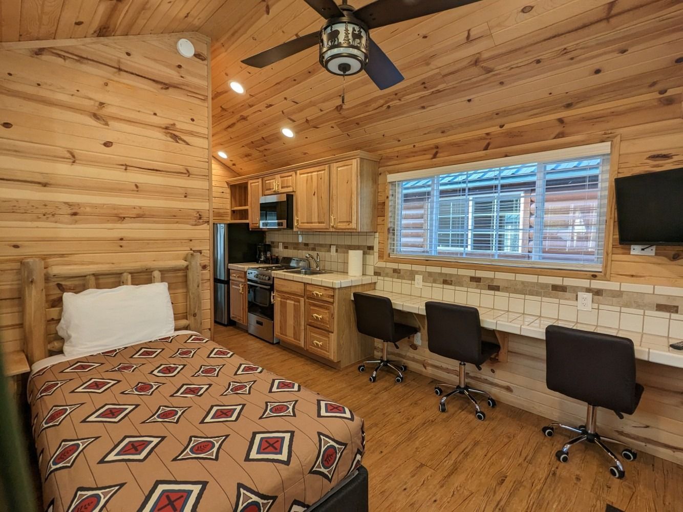 A bedroom in a log cabin with a bed , desk , chairs and a ceiling fan.