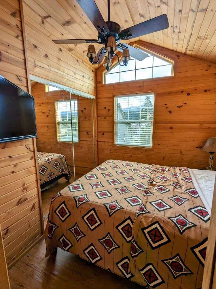 A bedroom in a log cabin with a bed , ceiling fan and television.
