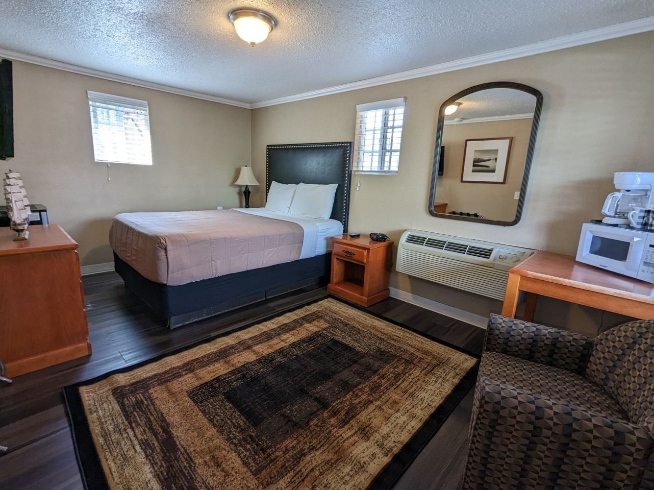 A hotel room with a king size bed , rug , chair , microwave and mirror.