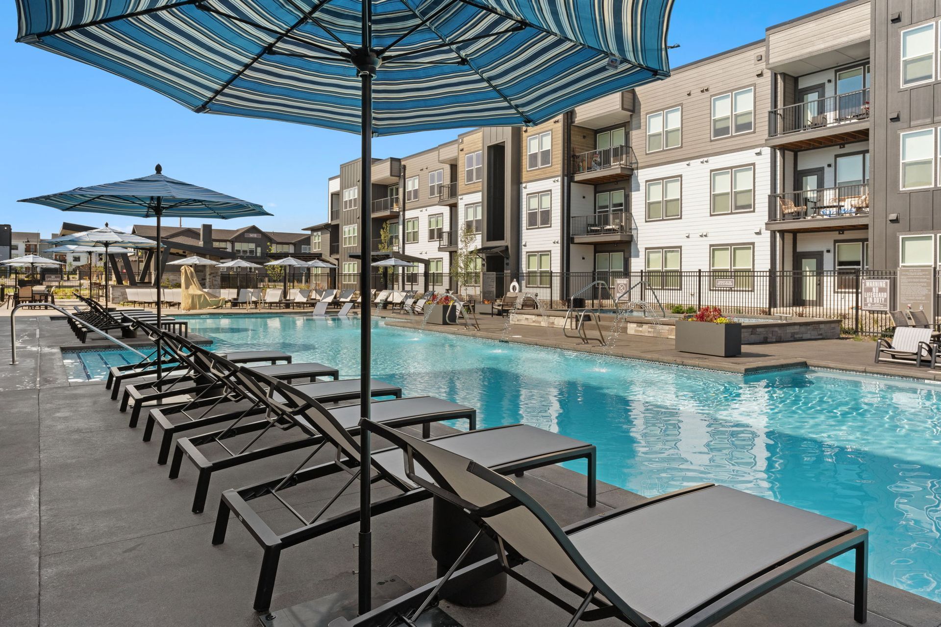 Poolside lounge chairs under umbrellas, in front of a modern apartment building with a pool.
