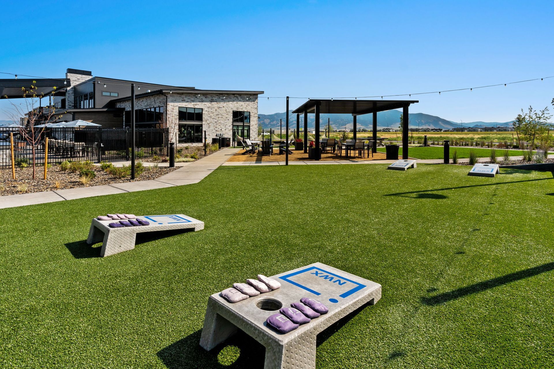 Cornhole game set up on green turf, outdoor area with modern building and pergola.