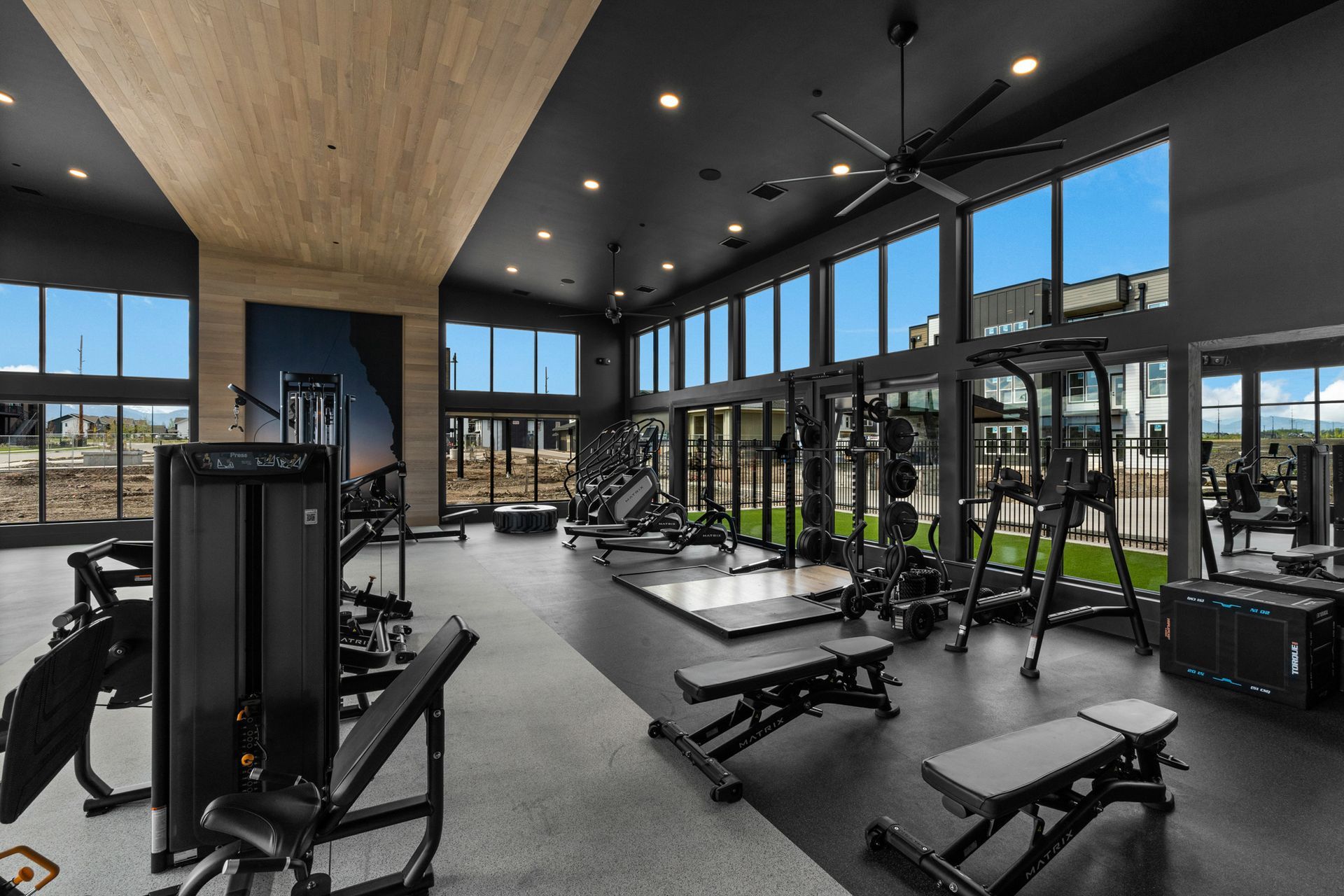 Gym interior with black equipment on a gray floor. Large windows offer an outside view.