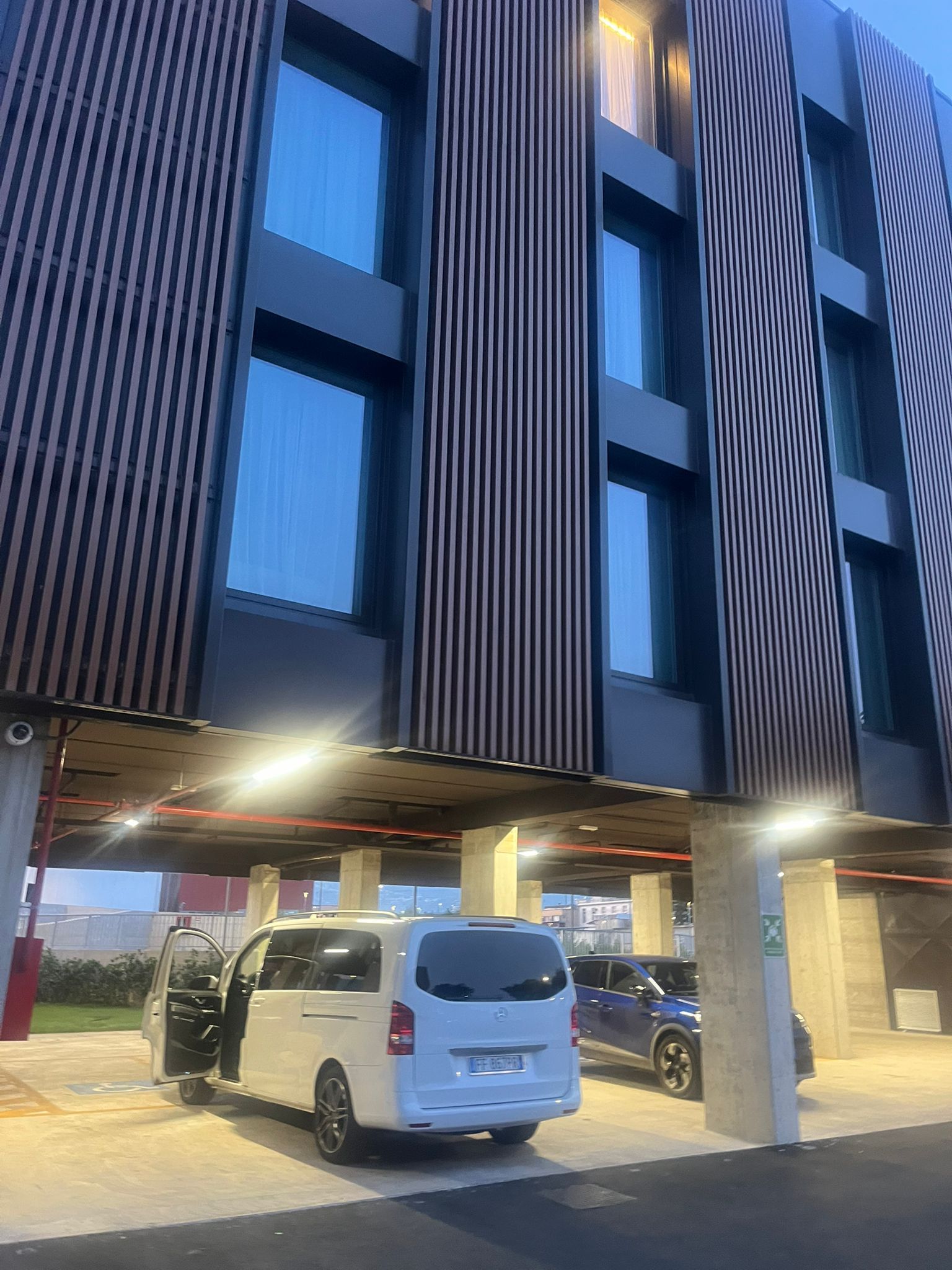 Edificio moderno con pannelli verticali in legno. Un furgone bianco e un'auto blu parcheggiati sotto una tettoia.