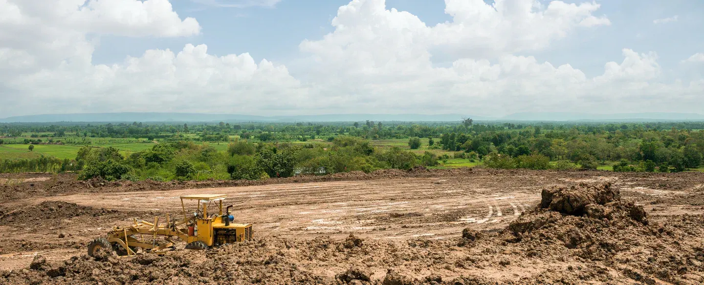 A bulldozer on a muddy field clearing land, with green trees and a cloudy sky in the background.