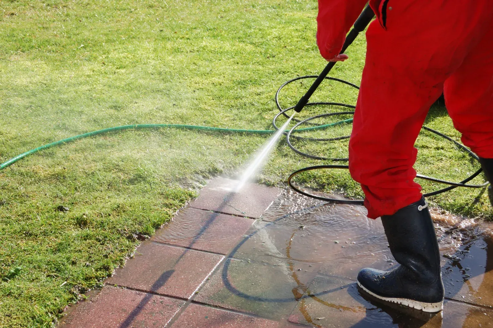 Person in red overalls power washing a brick patio next to grass.