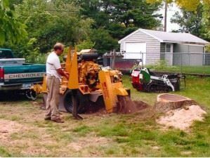 Man using a yellow backhoe to dig a trench beside a trailer in a grassy yard