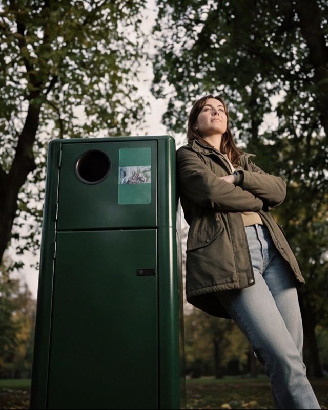 Person leaning against a Reverse vending machine in a park, arms crossed, looking upward.