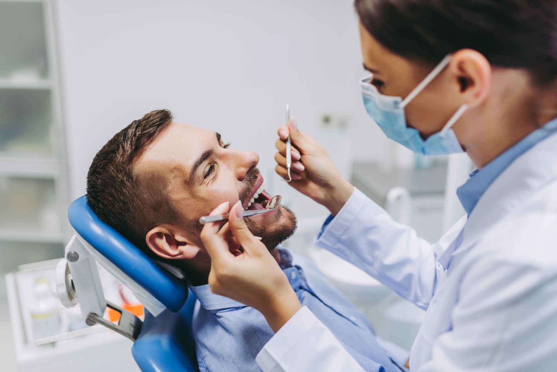 Dentist examining a patient's mouth with tools in a dental office. The patient is sitting in a blue chair.