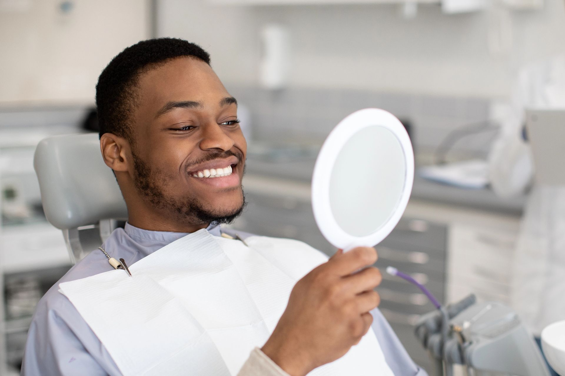 Man in a dental chair smiles, holding a mirror, looking at his teeth.