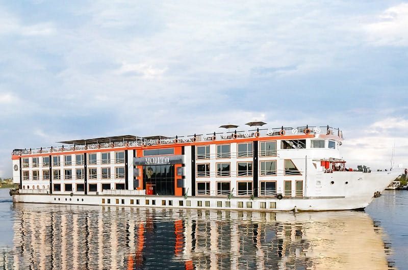 River cruise ship sailing on water. White and orange with many windows, reflecting in water.