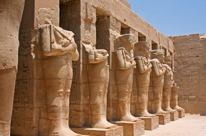 Row of ancient sandstone statues at the Karnak Temple in Egypt, with eroded heads and detailed garments.