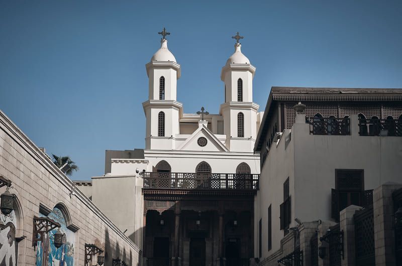 White church with two towers and crosses, viewed between buildings under a blue sky.