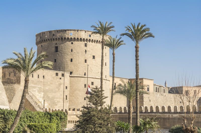 Stone fortress with tall cylindrical tower, palm trees in front, under a clear blue sky in Cairo, Egypt.
