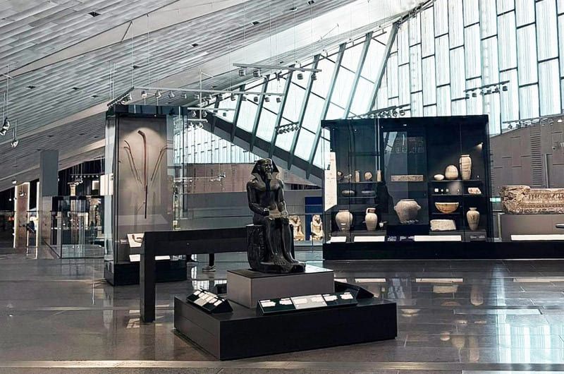 Museum interior with a seated statue in the foreground and glass display cases with artifacts.