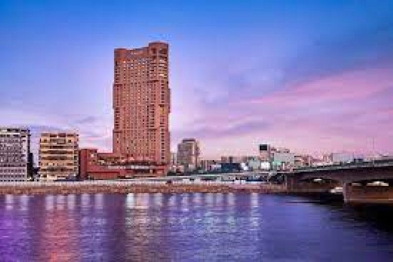 City skyline at dusk. Tall skyscraper, bridge, and calm water with reflections under a colorful sky.
