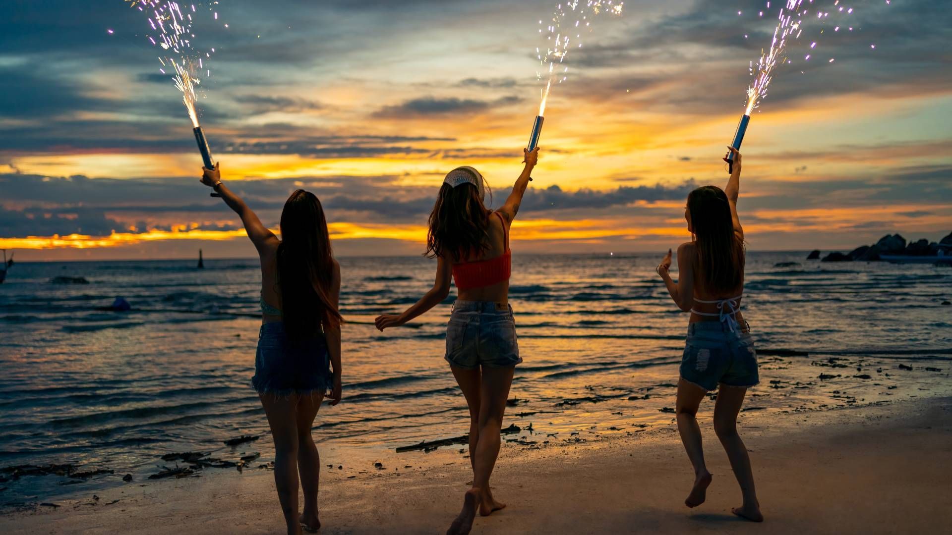 Three women are holding sparklers on the beach at sunset.
