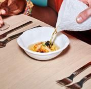 A person pours broth over food in a white bowl at a table set with utensils.