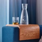 Wooden sofa arm tray holding a water carafe and a glass with liquid; blue backdrop.