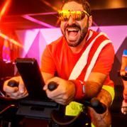 Man in sunglasses enthusiastically rides a stationary bike, red and yellow outfit, colorful gym setting.