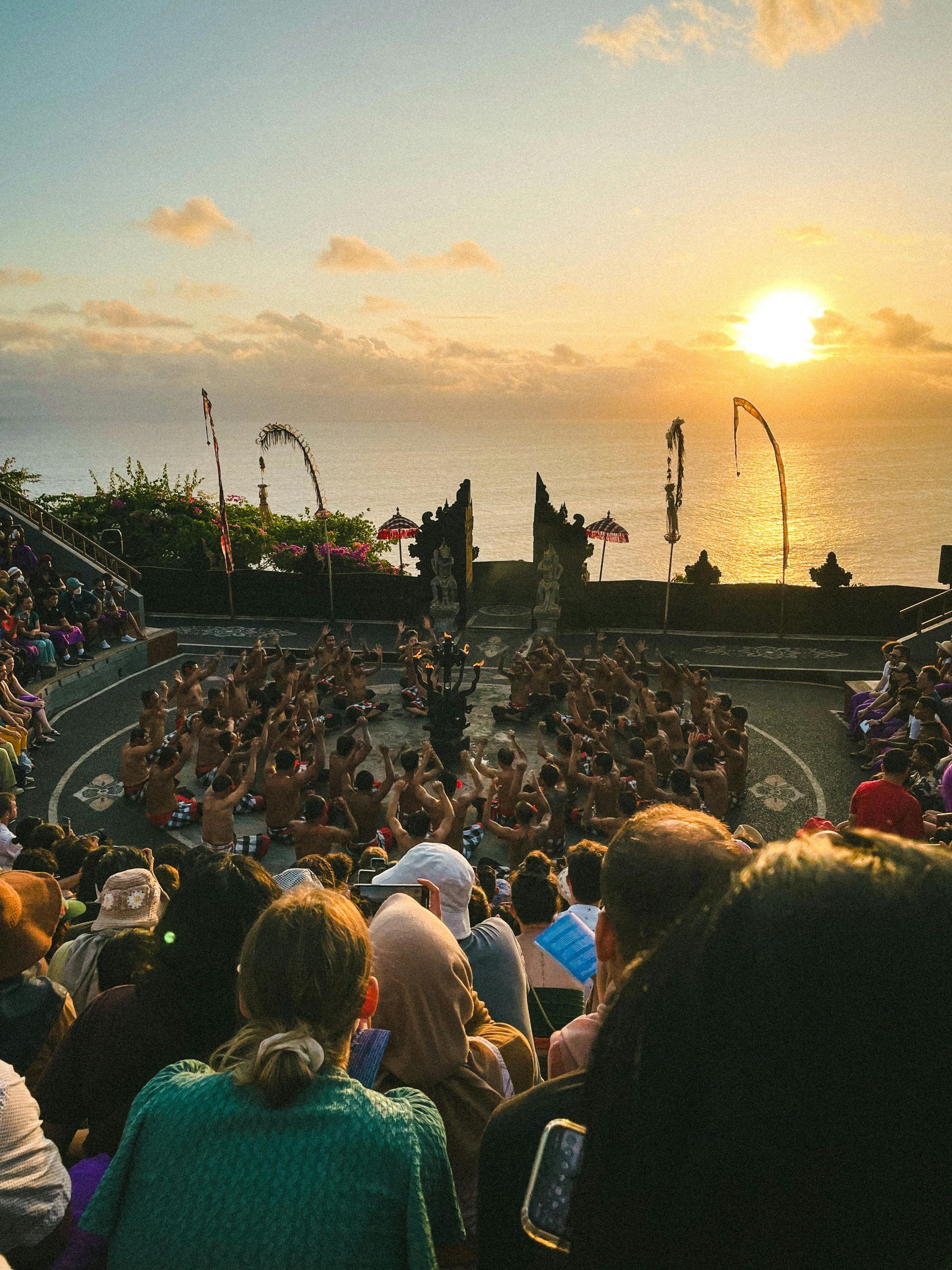 Sunset performance in Bali. People watch dancers in a circular arena near the ocean; golden light bathes the scene.