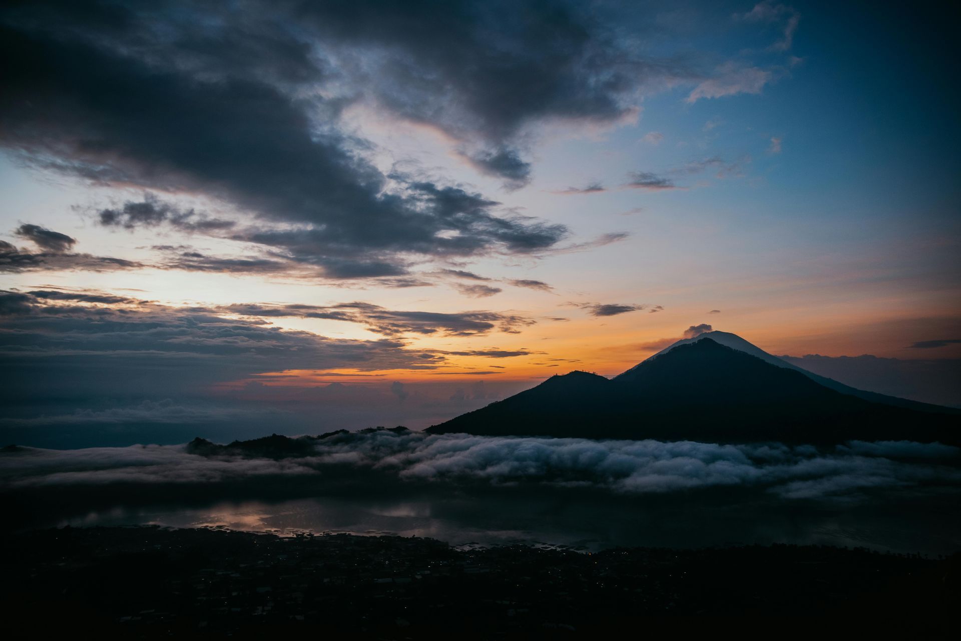 Mountain silhouettes against a sunrise sky; clouds blanket a lake below.