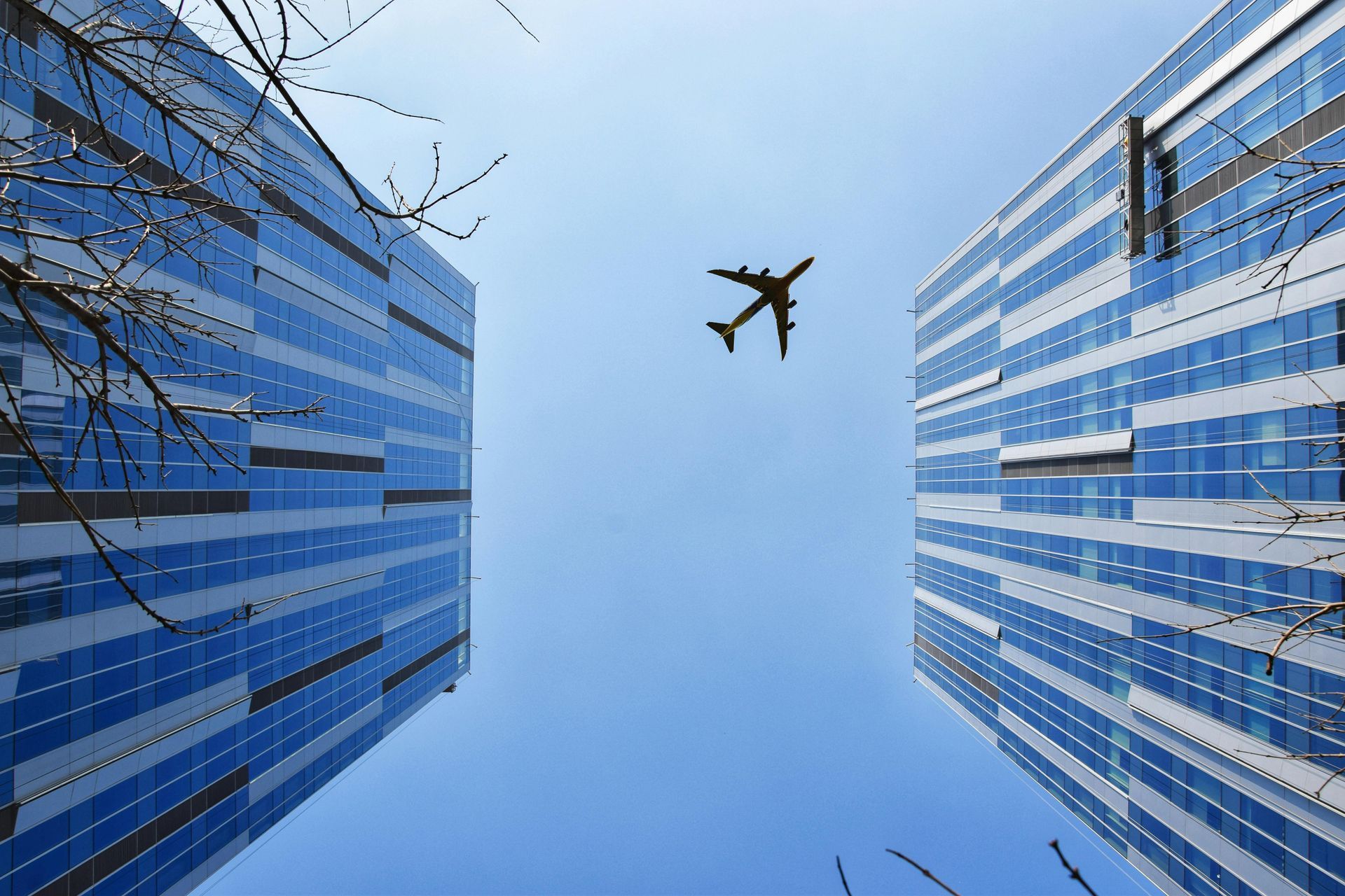 Airplane flying between two blue-glass skyscrapers under a clear sky.