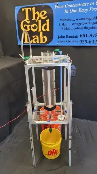 Small gold processing machine with pipes and buckets beside a “The Gold Lab” sign.