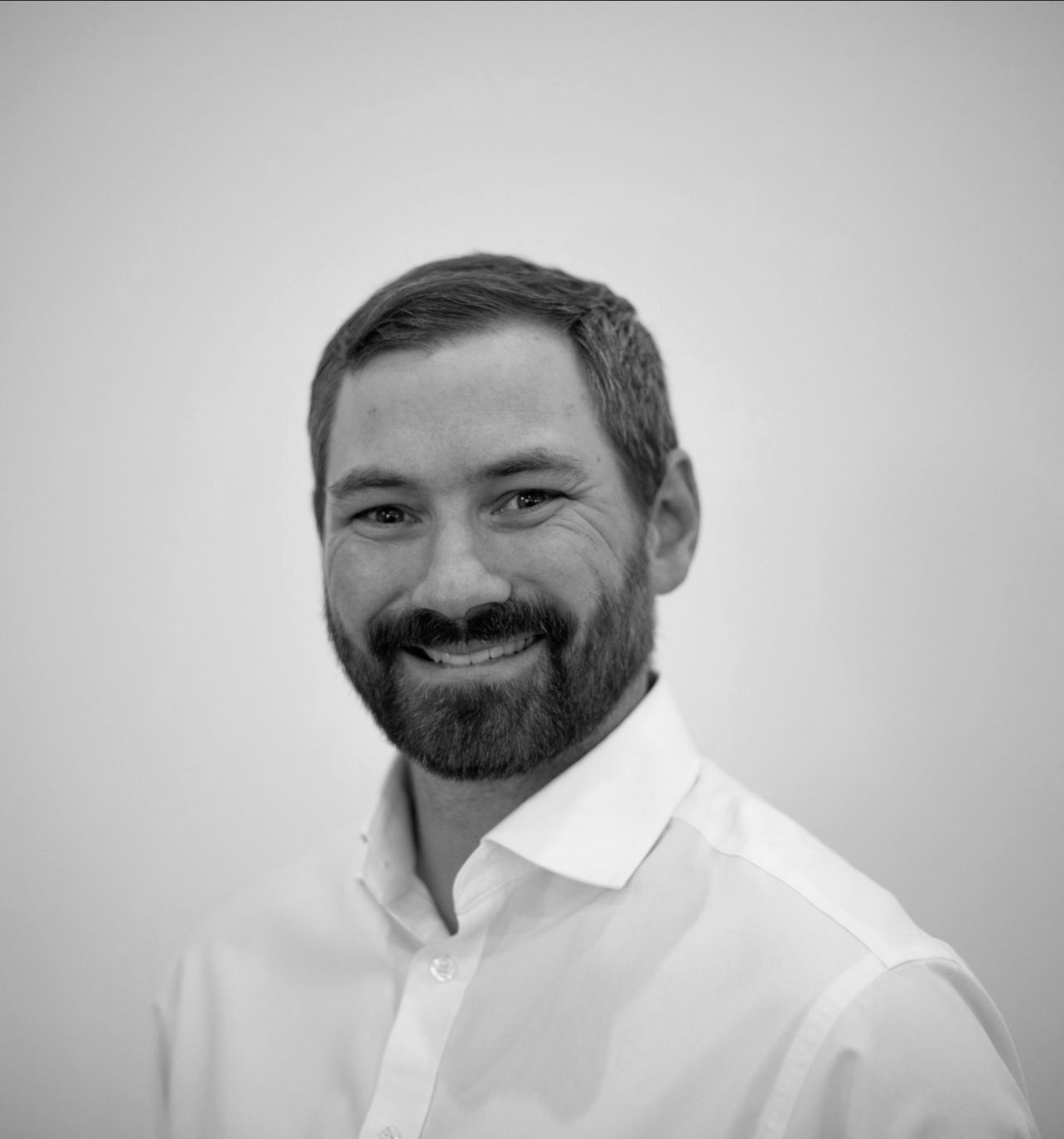 Man with a beard smiling, wearing a white collared shirt, headshot.