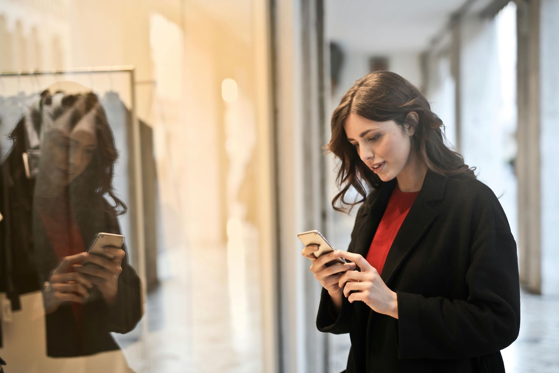 Woman in black coat looks at phone in front of store window, sunlight.
