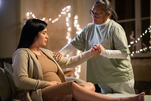 Pregnant woman with an older woman touching her arm, both indoors. Woman is sad.