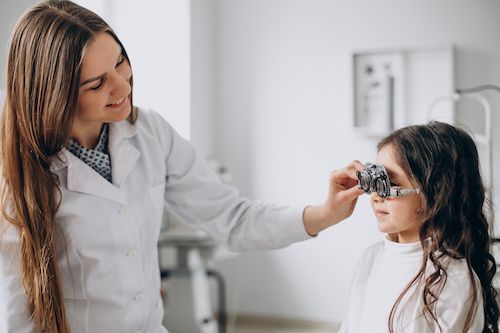 Optometrist examines a young girl's eyes with a phoropter in an examination room.